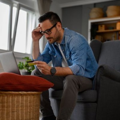 Man sitting at laptop with credit card in hand looking worried. Adobe.